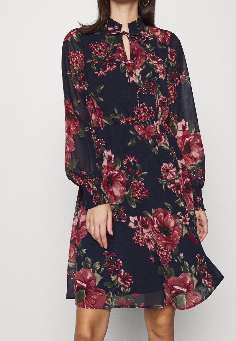 Femme portant une robe bleu marine à manches longues avec de grands motifs floraux rouges et roses, dotée d'un col montant avec une petite ouverture en forme de serrure et un lien.