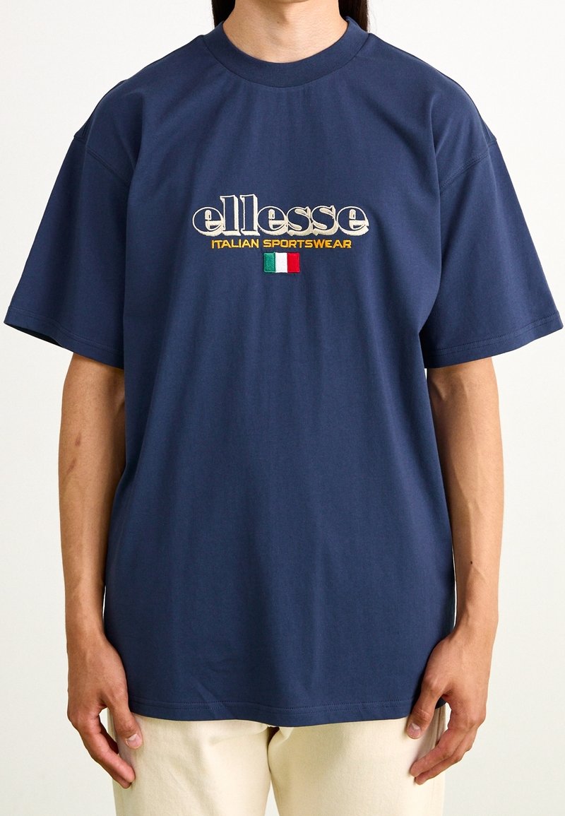 T-shirt en coton bleu marine à manches courtes, avec le logo "ellesse" et une broderie du drapeau italien au centre. Coupe décontractée et texture lisse.