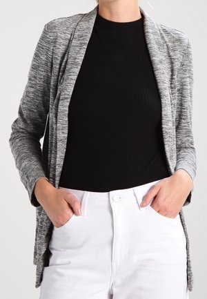 Kvinde iført en grå åben cardigan over en sort ribbet top med hvide højttaljede bukser, hænderne hviler i lommerne.