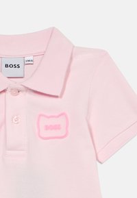 Ροζ πόλο πουκάμισο με υφασμάτινη υφή, διαθέτοντας άνοιγμα με κουμπιά και ανυψωμένη λαστιχένια ετικέτα λογότυπου σε αντίθετη απόχρωση.