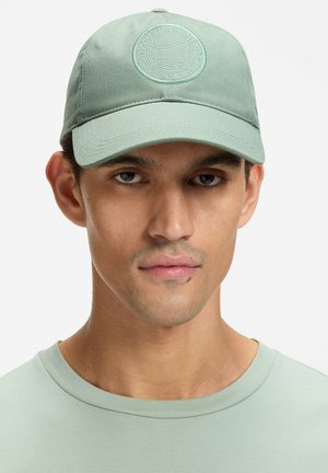 Green cotton cap with a circular embroidered logo on the front, flat brim, and adjustable strap at the back. Smooth texture.