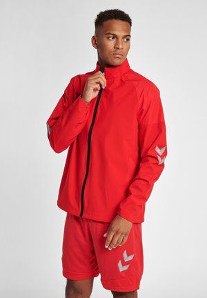Rote Sportjacke mit hohem Kragen und durchgehendem Reißverschluss. Mit reflektierenden grauen Akzenten an den Ärmeln. Trägt dazu passende rote Shorts.