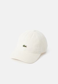 Cappellino da baseball in corduroy bianco con visiera curva. Presenta un piccolo logo di coccodrillo verde sul davanti. Superficie testurizzata e dettagli di cucitura.