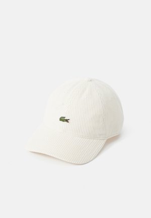 Cappellino da baseball in corduroy bianco con visiera curva. Presenta un piccolo logo di coccodrillo verde sul davanti. Superficie testurizzata e dettagli di cucitura.