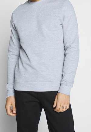 Mann trägt ein hellgraues Langarm-Sweatshirt und schwarze Hose, mit Tätowierungen an den Fingern seiner rechten Hand sichtbar.