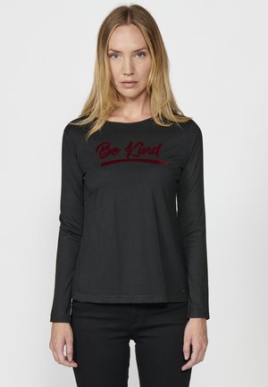 T-shirt noir à manches longues avec un col rond comportant un texte en velours "Bo Kind" sur la poitrine. Texture lisse, coupe décontractée.