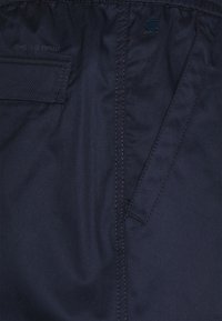 Tmavě modrá látka, ukazující švy a detaily kapsy. Obsahuje logo G-Star RAW v decentním designu. Hladká textura, polyestrová směs.