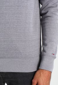 Suéter gris en mezcla de algodón, con puños y dobladillo acanalados. Un pequeño acento de logotipo en rojo, blanco y azul en el puño izquierdo.