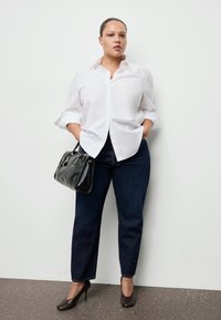 Žena stojící u bílé stěny, oblečená v bílé košili, tmavě modrých džínách a černých podpatcích, držící v ruce černou kabelku s rukama v kapsách.