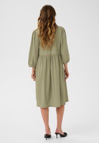 Grünes, strukturiertes Midikleid mit puffärmel und taillierter Taille. Der glatte Stoff reicht bis zur Wade und wird mit schwarzen, hohen Mules kombiniert.