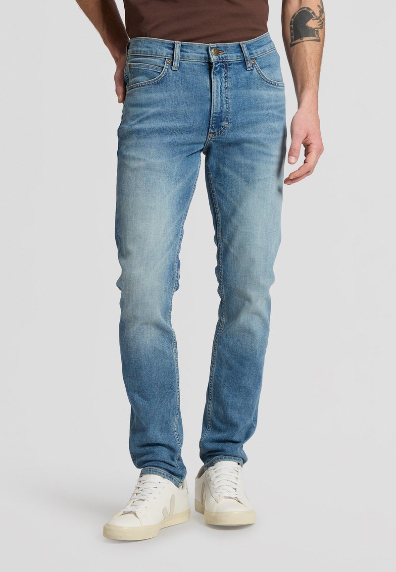 Homme portant un jean bleu délavé, des baskets blanches et une chemise marron, debout avec une main dans la poche sur un fond uni.