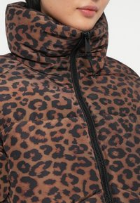 Tuvu attēls ar personu, kas valkā brūnu un melnu leoparda raksta puffer džemperi ar augstu apkakli un melnu rāvējslēdzēju.