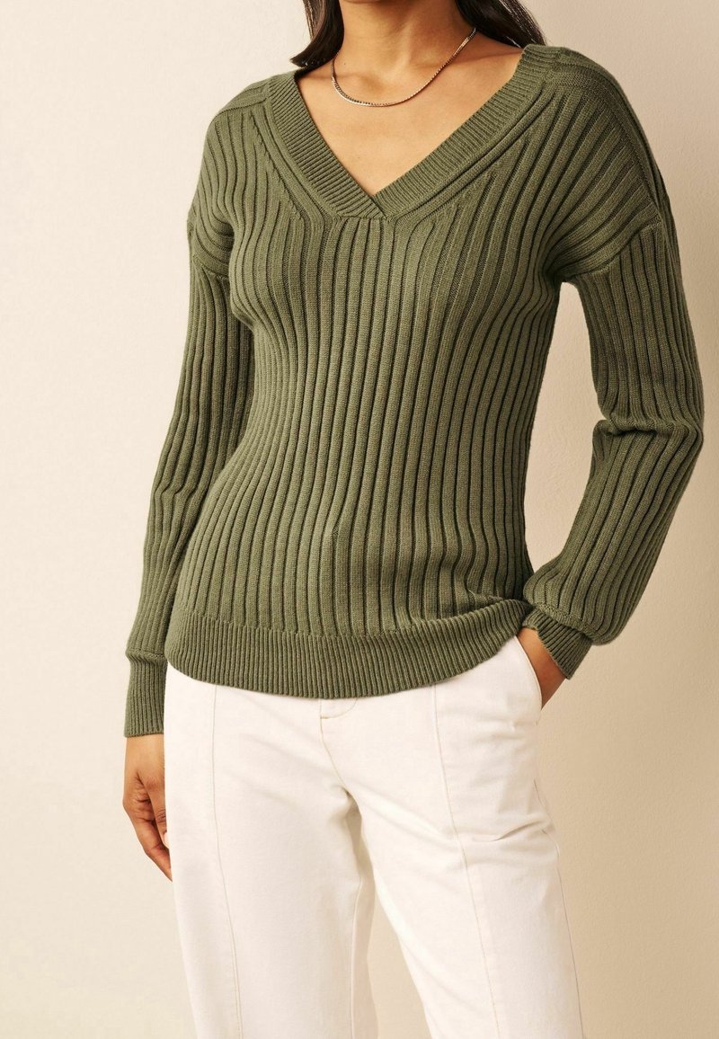 Femme portant un pull en maille côtelée vert olive à col en V et un pantalon blanc, avec une main dans la poche, sur un fond clair.