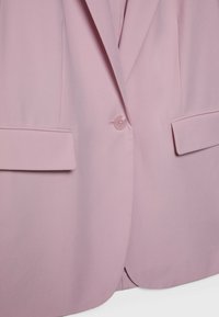 Blazer rose clair avec une fermeture à un bouton et deux poches à rabat à l'avant, présenté de la taille à la moitié de la poitrine.