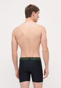Schwarze Boxershorts aus glattem Material mit einem breiten, elastischen Bund in Neon-Grün, auf dem "UNDER" auffällig gedruckt ist.