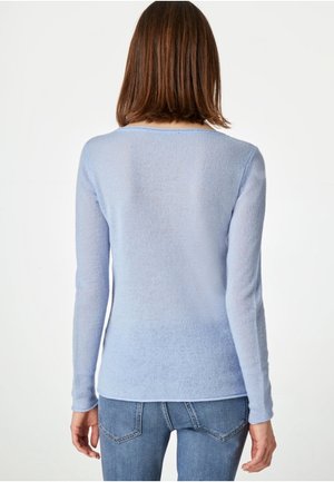 Femme aux cheveux bruns et raides portant un pull bleu clair à manches longues et un jean bleu, de dos sur un fond blanc.