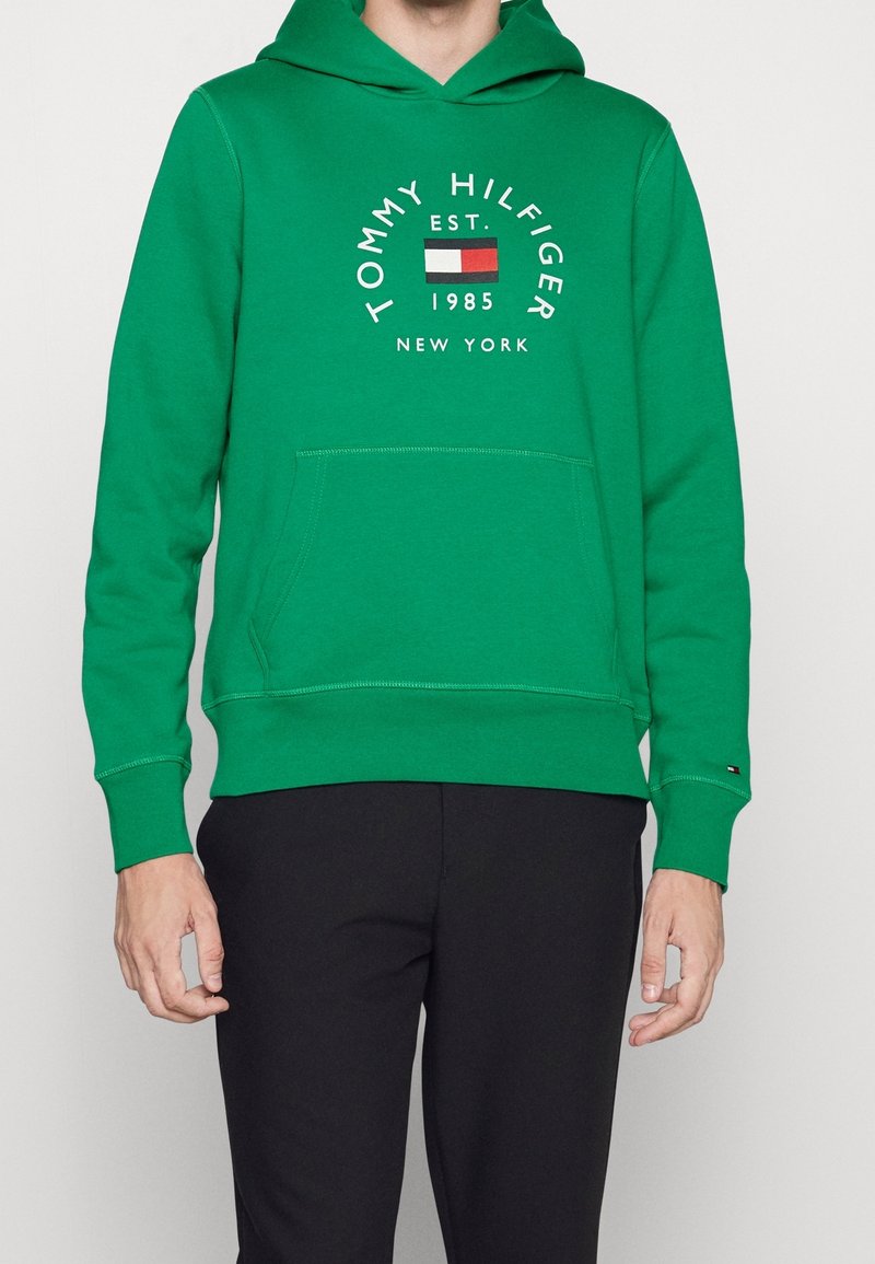 Personne portant un sweat à capuche vert avec le texte et le logo « Tommy Hilfiger Est. 1985 New York », associé à un pantalon noir, debout sur un fond blanc.