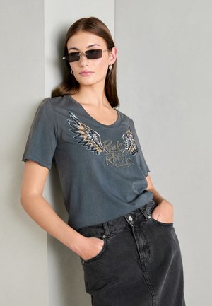 T-shirt gris à manches courtes avec un graphisme d'ailes et le texte "Rock & Roll". Associé à une jupe en denim noire et accessoirisé avec des boucles d'oreilles en argent et des lunettes de soleil.