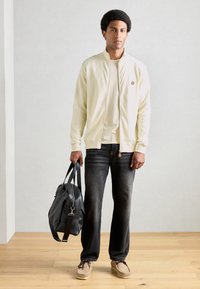 Lys cremefarvet zip-up jakke med ribbede manchetter, beige t-shirt, mørke jeans og grå sko. Sort lædertaske med håndtag og skulderrem.