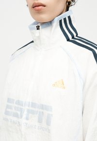 Άτομο που φοράει λευκό αθλητικό μπουφάν Adidas με μαύρες ρίγες στους ώμους και χρυσό λογότυπο Adidas στο στήθος, φερμουάρ κλειστό μέχρι τον λαιμό.