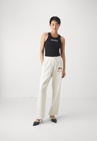 Čierny rebrovaný top na ramienka s logom "FIORUCCI", v kombinácii s krémovými teplákovými nohavicami s vyšívaným vzorom. Model nosí čierne lodičky.