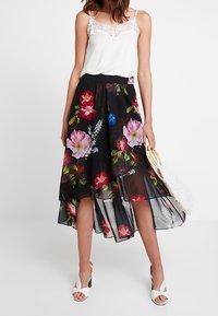 Falda negra con flores que presenta un diseño asimétrico con patrones de flores coloridas, combinada con una camiseta sin mangas con encaje blanco y zapatos blancos.