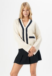 Femeie blondă îmbrăcată într-o jachetă de culoare crem cu trim și nasturi negri, asortată cu o fustă mini plisată neagră, pozând pe un fundal alb.