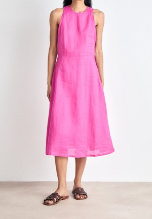 Žena v bezrukávých jasně růžových midi šatech a hnědých nízkých nazouvacích sandálech stojí před světlou zdí.