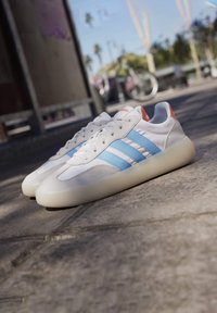 Vita sneakers med blå ränder, material av mocka och läder, rundad tå, gummisula och orangea detaljer nära hälen.