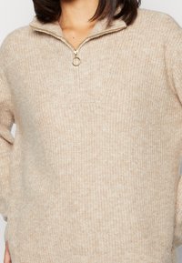 Beige, gestrickter Pullover mit einem halben Reißverschluss und goldenen Hardware-Akzenten. Strukturiertes Material mit einem lässigen Schnitt und gerippten Details.