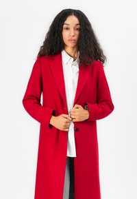 Manteau en laine rouge au design ajusté, avec des revers crantés et des manches longues. Il présente une fermeture à boutons sur le devant et est porté par-dessus une chemise blanche.