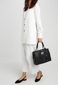 Sac en cuir noir avec des accents cloutés, des garnitures en argent et des poignées tressées. Porté avec un costume à rayures blanches et des ballerines noires.