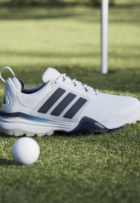 Λευκό παπούτσι του γκολφ με μαύρες ρίγες δίπλα σε μια μπάλα του γκολφ σε πράσινο γρασίδι κοντά σε σημαία σε γήπεδο γκολφ.