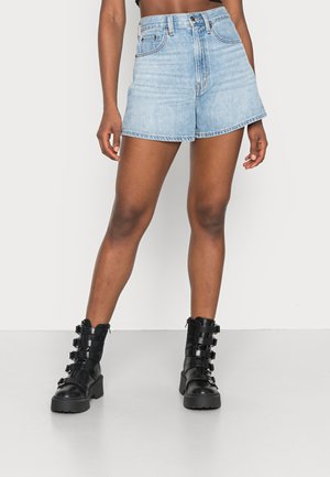 Personne portant un short en denim bleu clair et des bottes de combat noires à boucle, debout devant un fond clair uni.