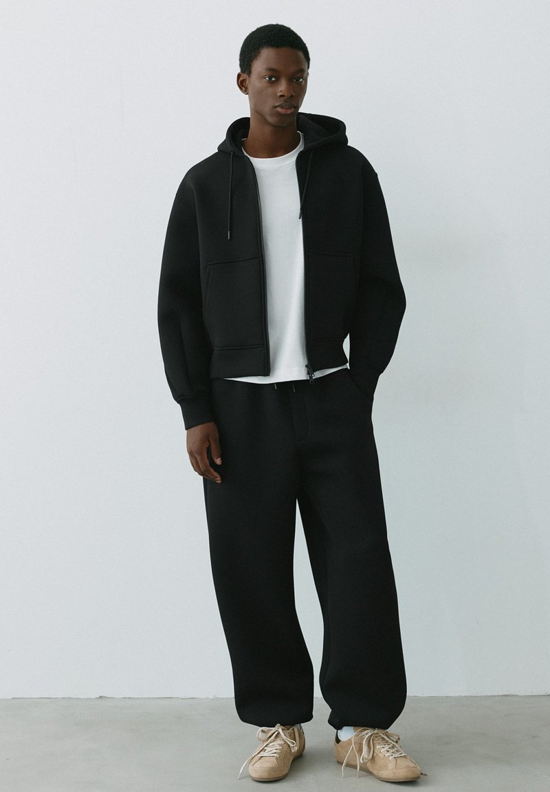 Jeune homme portant un sweat à capuche noir zippé, un t-shirt blanc, un pantalon noir ample et des baskets beiges, debout contre un mur clair uni.