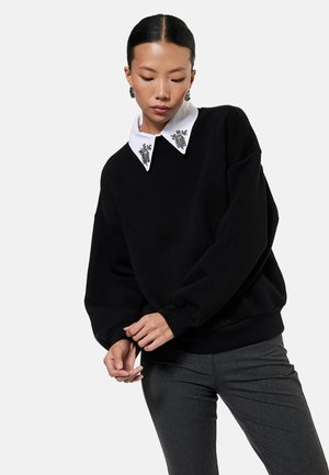 Vrouw draagt een zwarte trui met een versierde witte kraag en grijze broek, poseert met handen gevouwen en hoofd naar beneden gekanteld.