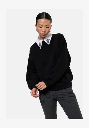 Mujer que lleva un suéter negro con un cuello blanco adornado y pantalones grises, posando con las manos entrelazadas y la cabeza inclinada hacia abajo.