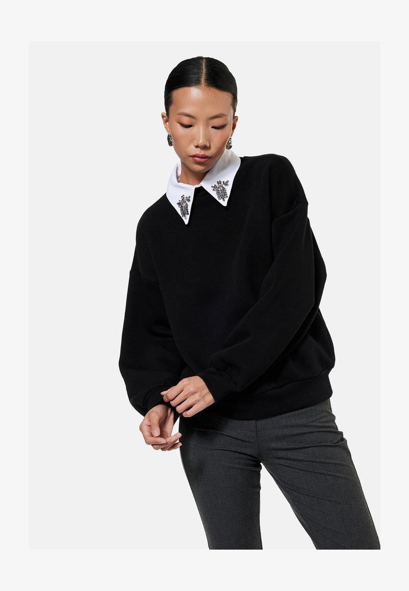 Frau trägt einen schwarzen Pullover mit verziertem weißem Kragen und grauer Hose, posiert mit verschränkten Händen und gesenktem Kopf.