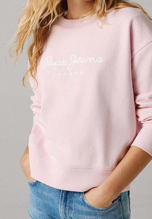 Personne portant un sweat Pepe Jeans London rose clair avec les mains dans les poches d’un jean bleu, sur un fond gris uni.