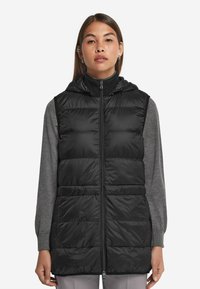 Czarna kamizelka puchowa z kapturem, z pikowanym wzorem, zamkiem z przodu i kieszeniami po bokach, noszona na szarym longsleeve.