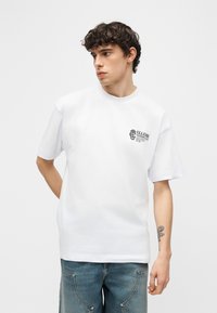 Νεαρός άνδρας με κατσαρά μαλλιά, φοράει λευκό κοντομάνικο πουκάμισο με λογότυπο στο στήθος και μπλε τζιν, στέκεται μπροστά σε απλό φόντο.