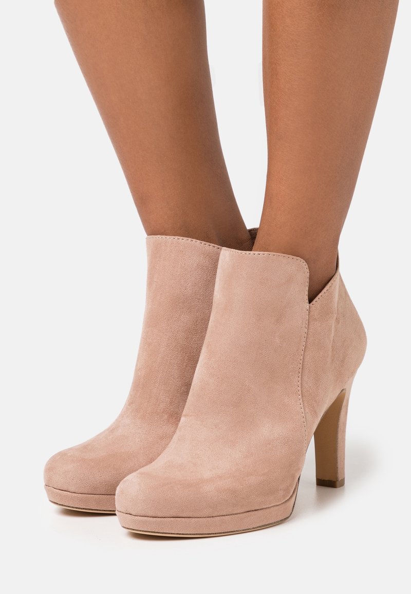 Bottines en daim beige clair à talons hauts portées aux pieds sur un fond uni.