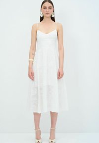 Witte midi-jurk met bloemmotief van kant, dunne bandjes en een nauwsluitend lijfje. Aangevuld met een vloeiende rok, gecompleteerd met lichtgekleurde hakken.