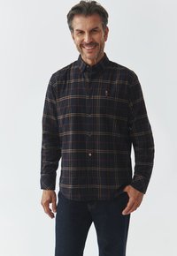 Cămașă în carouri închise, cu linii albastre, roșii și galbene, guler tip button-down, mâneci lungi și un mic logo pe pieptul stâng. Purtați cu blugi închiși la culoare.