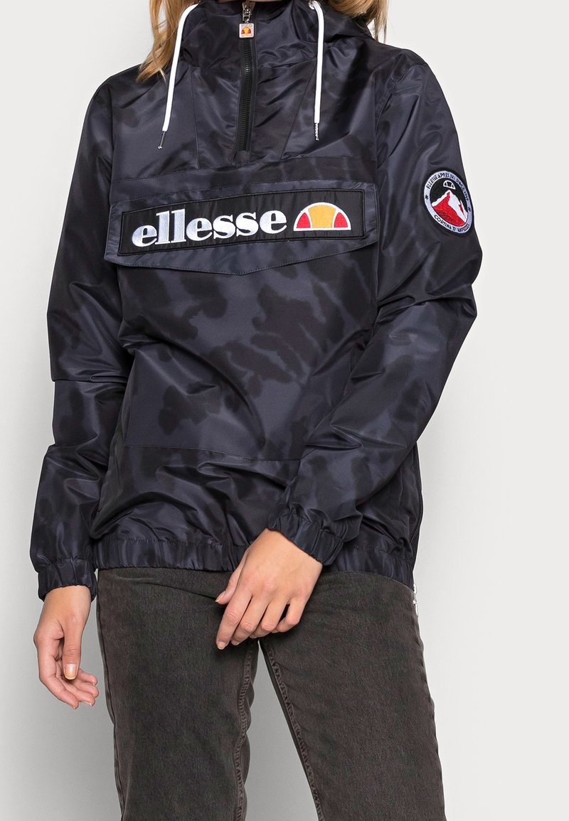 Svart vindjacka med halvdragkedja, stor framficka, elastiska manschetter och "ellesse"-logotyp. Har ett subtilt mönster och huva.