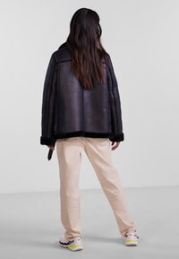 Black leather jacket with textured finish, oversized design, and fur trim at cuffs. Paired with beige pants and white sneakers.