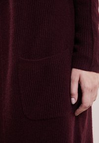 Cardigan in magenta lavorato a maglia con texture a coste, maniche lunghe e una tasca frontale sul lato sinistro vicino all'orlo.