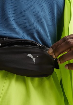 Sacoche noire avec fermeture éclair, arborant un logo Puma argenté, portée sur une chemise bleue texturée et une couche extérieure vert fluo.