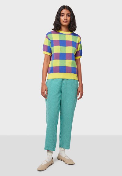 Top de punto de manga corta con un patrón de cuadros multicolores en amarillo, azul y púrpura; combinado con pantalones holgados a rayas verdes y zapatos beige.