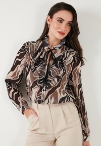 Blusa trasparente marrone e beige con motivo a rouches, dettaglio in nero al collo e maniche lunghe, abbinata a pantaloni su misura di colore chiaro.